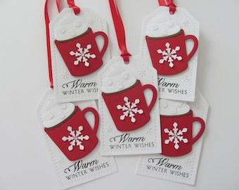 May include: White gift tags with a red mug design, topped with white whipped cream and a snowflake. The tags have the words "Warm Winter Wishes" and are tied with red ribbon. A festive holiday theme.