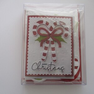 K&ouml;nnte beinhalten: Eine transparente Plastikbox mit Weihnachtskarten. Die Vorderseite der Karte zeigt ein Zuckerstangen-Design mit einer roten Schleife und dem Schriftzug "Merry Christmas". Die Karten haben einen roten, gezackten Rand.