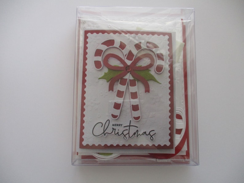 K&ouml;nnte beinhalten: Eine transparente Plastikbox mit Weihnachtskarten. Die Vorderseite der Karte zeigt ein Zuckerstangen-Design mit einer roten Schleife und dem Schriftzug "Merry Christmas". Die Karten haben einen roten, gezackten Rand.