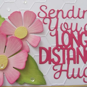 May include: A white card with a pink and green floral design. The card has the text "Sending you a long distance hug" in pink.