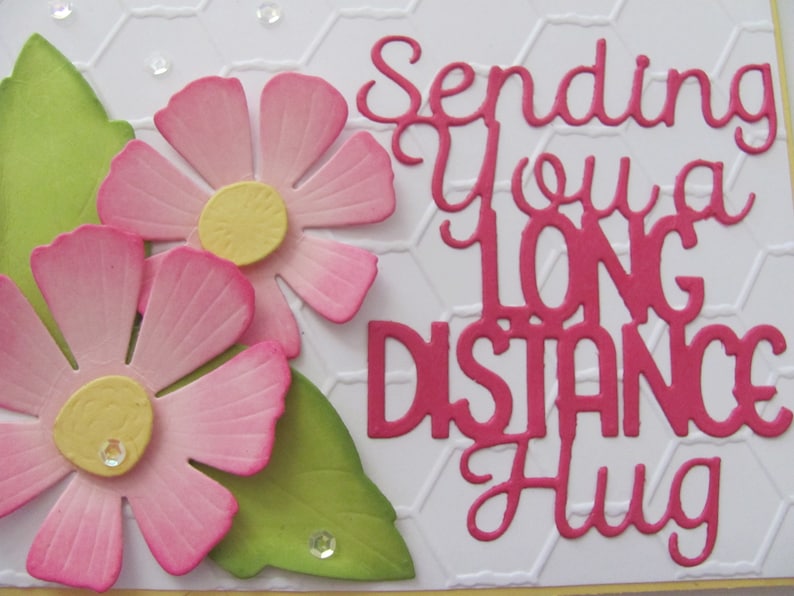 May include: A white card with a pink and green floral design. The card has the text "Sending you a long distance hug" in pink.