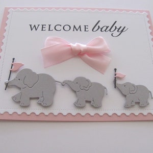 K&ouml;nnte beinhalten: Eine rosa und wei&szlig;e Babyparty-Karte mit dem Text "WELCOME baby" und drei grauen Elefanten, die rosa Flaggen halten.