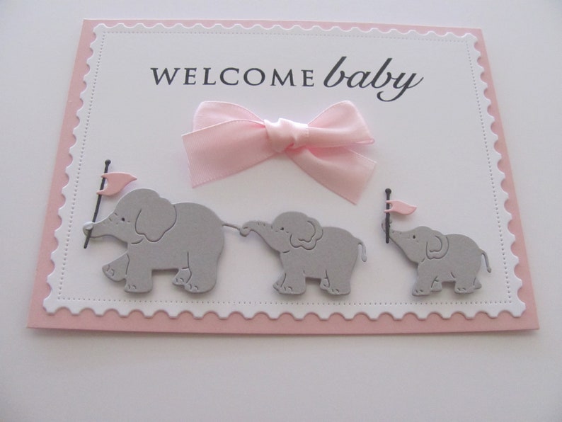 K&ouml;nnte beinhalten: Eine rosa und wei&szlig;e Babyparty-Karte mit dem Text "WELCOME baby" und drei grauen Elefanten, die rosa Flaggen halten.