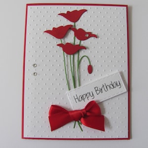 May include: A white card with a red border and a red bow. The card has a white background with a dotted pattern. The card has a bouquet of red paper flowers and the text "Happy Birthday".