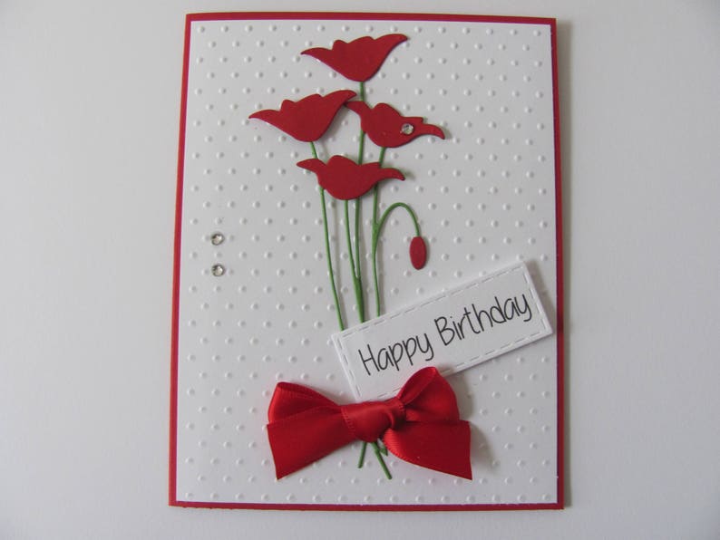 May include: A white card with a red border and a red bow. The card has a white background with a dotted pattern. The card has a bouquet of red paper flowers and the text "Happy Birthday".