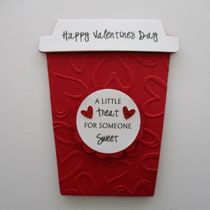 May include: A red Valentine's Day-themed coffee cup-shaped decoration. The cup has a white top with "Happy Valentines Day" printed on it. A white circle with the text "A little treat for someone sweet" and two red glitter hearts is in the center.