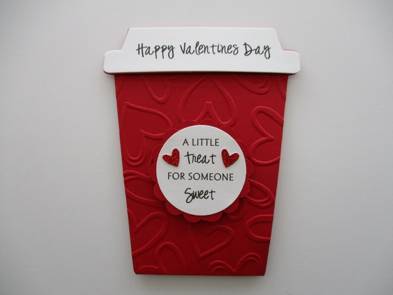 May include: A red Valentine's Day-themed coffee cup-shaped decoration. The cup has a white top with "Happy Valentines Day" printed on it. A white circle with the text "A little treat for someone sweet" and two red glitter hearts is in the center.