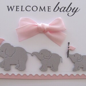 K&ouml;nnte beinhalten: Eine wei&szlig;e Karte mit rosa gewellten R&auml;ndern und einer rosa Schleife. Die Karte sagt "WELCOME baby" in schwarzen Buchstaben. Drei graue Papierelefanten mit rosa Flaggen befinden sich auf der Karte.