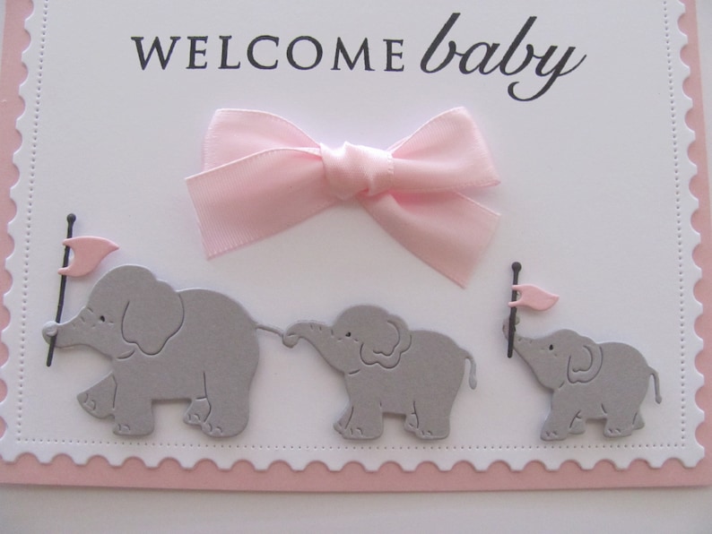 K&ouml;nnte beinhalten: Eine wei&szlig;e Karte mit rosa gewellten R&auml;ndern und einer rosa Schleife. Die Karte sagt "WELCOME baby" in schwarzen Buchstaben. Drei graue Papierelefanten mit rosa Flaggen befinden sich auf der Karte.
