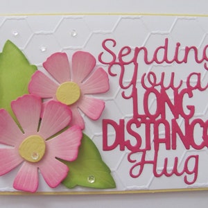 May include: A white card with a pink and green floral design. The card has the text "Sending you a long distance hug" in pink.