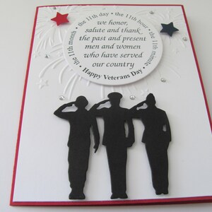 Pu&ograve; includere: Un biglietto fatto a mano per il Veterans Day con un bordo rosso. La carta bianca presenta un testo circolare che recita "Happy Veterans Day" e una silhouette di tre figure che salutano. Sono inclusi abbellimenti a forma di stella rossa e blu.