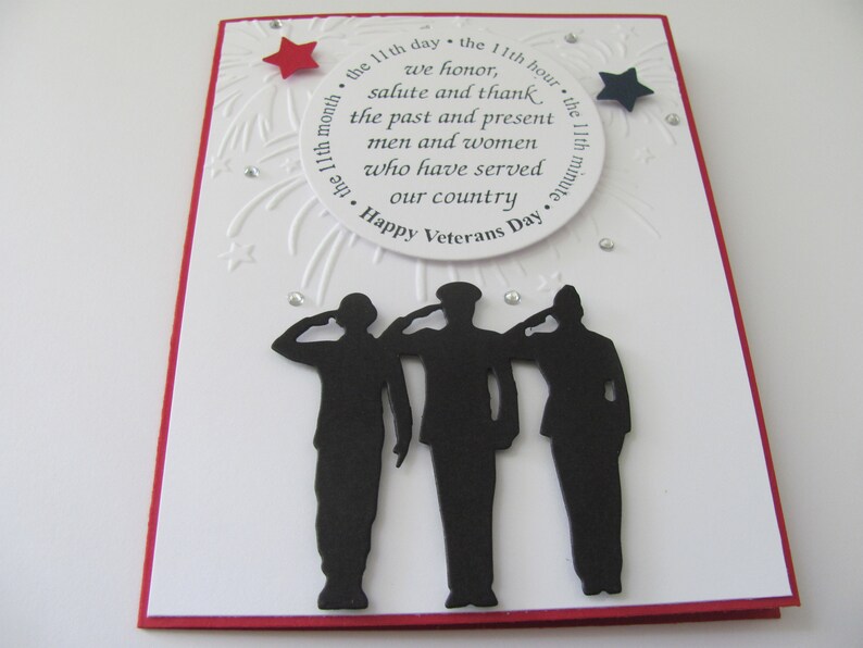 Pu&ograve; includere: Un biglietto fatto a mano per il Veterans Day con un bordo rosso. La carta bianca presenta un testo circolare che recita "Happy Veterans Day" e una silhouette di tre figure che salutano. Sono inclusi abbellimenti a forma di stella rossa e blu.
