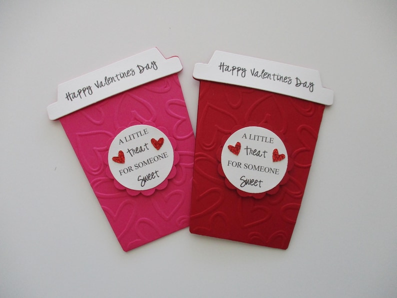 May include: Two Valentine's Day-themed coffee cup-shaped cards. One is pink, the other red, both with embossed heart patterns. Each has a white label with the text "A LITTLE treat FOR SOMEONE Sweet" and "Happy Valentines Day".