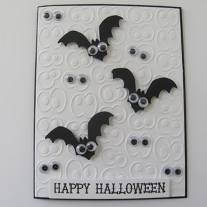 May include: A rectangular greeting card with a white embossed background featuring repeating eye patterns. Three black paper bats with googly eyes are attached to the front. Several loose googly eyes are scattered across the card. The text HAPPY HALLOWEEN is printed in black on a white strip at the bottom.