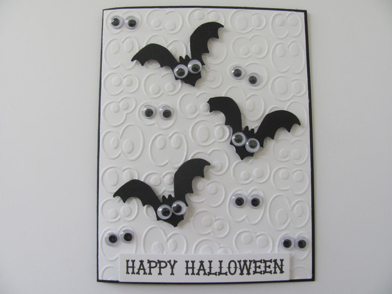 May include: A rectangular greeting card with a white embossed background featuring repeating eye patterns. Three black paper bats with googly eyes are attached to the front. Several loose googly eyes are scattered across the card. The text HAPPY HALLOWEEN is printed in black on a white strip at the bottom.
