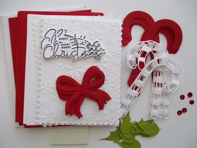 K&ouml;nnte beinhalten: Ein festliches Weihnachtskartenset mit wei&szlig;em Pr&auml;gedesign, roter Schleife und dem Schriftzug "Merry Christmas". Enth&auml;lt rote Umschl&auml;ge, Zuckerstangen-Dekorationen, gr&uuml;ne Stechpalmenbl&auml;tter und rote Pailletten.