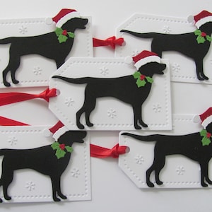 Pu&ograve; includere: Cinque etichette regalo bianche con immagini di silhouette nere di cani che indossano cappelli di Babbo Natale e agrifoglio. Le etichette hanno un bordo punteggiato e un motivo a fiocco di neve.