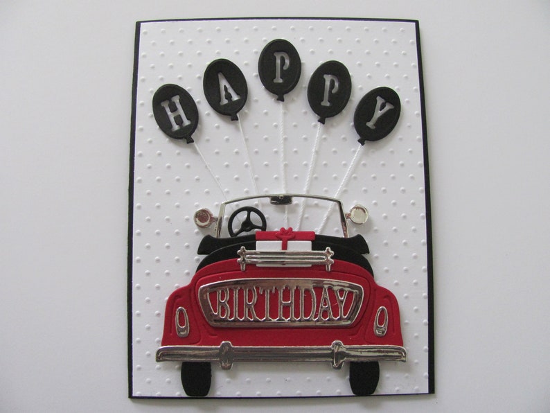 May include: A red vintage car with black wheels and silver trim. The car has a white gift box in the back and black balloons with silver letters spelling "HAPPY" above it. The car has a silver number plate that says "BIRTHDAY".