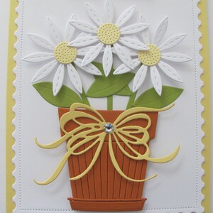 May include: A white card with a yellow scalloped border. The card features a paper craft design of three white daisies with yellow centers in a brown pot with a yellow bow.