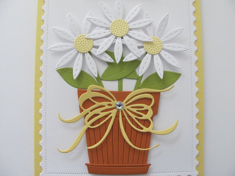 May include: A white card with a yellow scalloped border. The card features a paper craft design of three white daisies with yellow centers in a brown pot with a yellow bow.