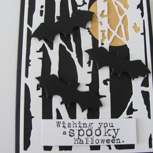 Puede incluir: Una tarjeta de felicitaci&oacute;n rectangular con fondo negro, una superposici&oacute;n blanca troquelada de siluetas de &aacute;rboles desnudos y una luna amarilla. Tres murci&eacute;lagos de papel negro est&aacute;n superpuestos. Una etiqueta blanca inferior dice Wishing you a spooky halloween.