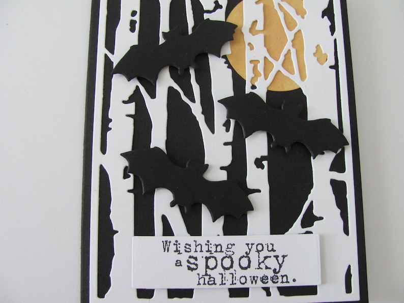 Puede incluir: Una tarjeta de felicitaci&oacute;n rectangular con fondo negro, una superposici&oacute;n blanca troquelada de siluetas de &aacute;rboles desnudos y una luna amarilla. Tres murci&eacute;lagos de papel negro est&aacute;n superpuestos. Una etiqueta blanca inferior dice Wishing you a spooky halloween.