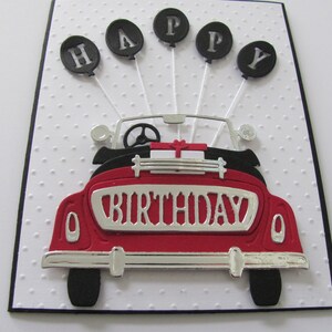 May include: A red vintage car with black wheels and a silver "Happy Birthday" sign on the back. The car has black balloons with white letters spelling out "Happy" above it.