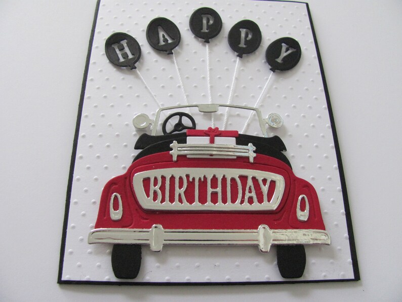 May include: A red vintage car with black wheels and a silver "Happy Birthday" sign on the back. The car has black balloons with white letters spelling out "Happy" above it.