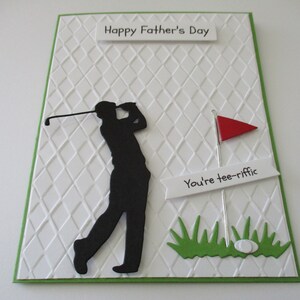 May include: A white card with a green border and a diamond pattern. The card has a black silhouette of a golfer swinging a club. There is a red flag and a white sign that says "You're tee-riffic". The card says "Happy Father's Day" at the top.