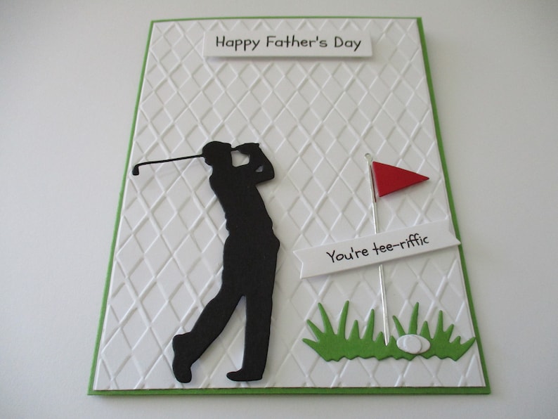 May include: A white card with a green border and a diamond pattern. The card has a black silhouette of a golfer swinging a club. There is a red flag and a white sign that says "You're tee-riffic". The card says "Happy Father's Day" at the top.