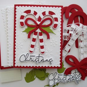 K&ouml;nnte beinhalten: Eine Sammlung von Bastelmaterialien zum Thema Weihnachten. Im Mittelpunkt steht eine wei&szlig;e Karte mit rotem Rand, die Zuckerstangen, eine rote Schleife und die Worte "Merry Christmas" zeigt. Weitere Elemente sind rote und wei&szlig;e Zuckerstangen, gr&uuml;ne Stechpalmenbl&auml;tter und rote Pailletten.