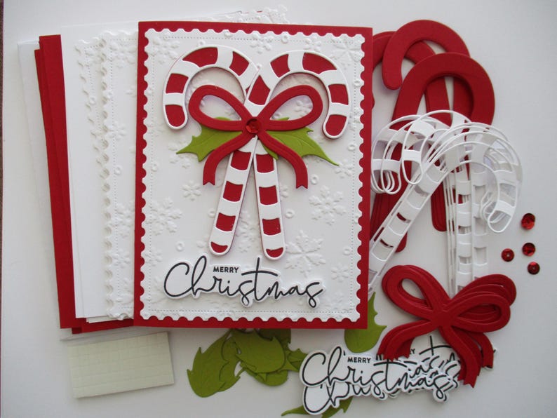 K&ouml;nnte beinhalten: Eine Sammlung von Bastelmaterialien zum Thema Weihnachten. Im Mittelpunkt steht eine wei&szlig;e Karte mit rotem Rand, die Zuckerstangen, eine rote Schleife und die Worte "Merry Christmas" zeigt. Weitere Elemente sind rote und wei&szlig;e Zuckerstangen, gr&uuml;ne Stechpalmenbl&auml;tter und rote Pailletten.