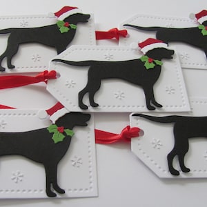 Pu&ograve; includere: Cinque etichette regalo bianche con una silhouette nera di un cane che indossa un cappello di Babbo Natale e agrifoglio. Le etichette hanno un bordo punteggiato e un motivo a fiocco di neve. Un nastro rosso &egrave; attaccato alle etichette.