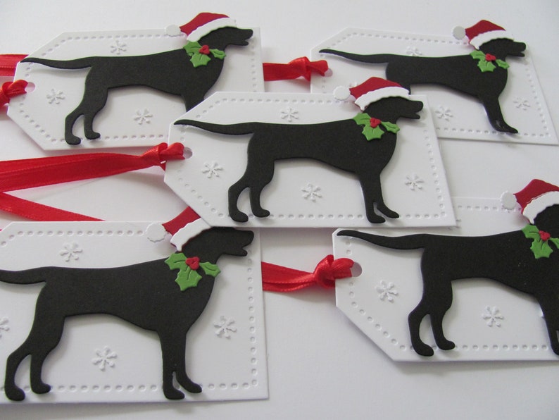 Pu&ograve; includere: Cinque etichette regalo bianche con una silhouette nera di un cane che indossa un cappello di Babbo Natale e agrifoglio. Le etichette hanno un bordo punteggiato e un motivo a fiocco di neve. Un nastro rosso &egrave; attaccato alle etichette.
