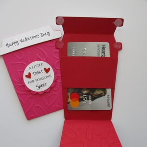May include: A pink paper coffee cup-shaped card with embossed hearts and the text "Happy Valentines Day." A red card holder with two credit cards is next to it. The card has the text "A little treat for someone sweet."