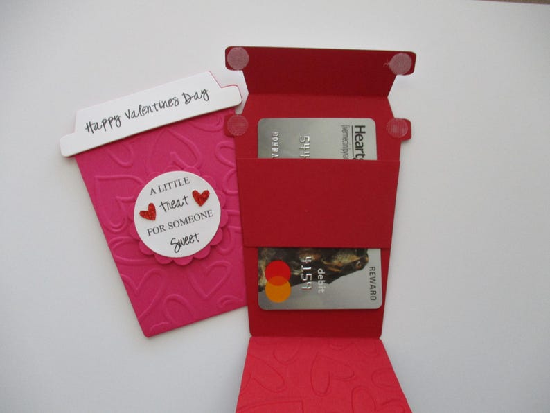 May include: A pink paper coffee cup-shaped card with embossed hearts and the text "Happy Valentines Day." A red card holder with two credit cards is next to it. The card has the text "A little treat for someone sweet."