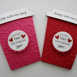 May include: Two Valentine's Day cards shaped like coffee cups. One is pink, the other red, both with embossed heart patterns. Each card has a white label with the text "A LITTLE TREAT FOR SOMEONE SWEET" and two red glitter hearts. The top reads "Happy Valentine's Day."