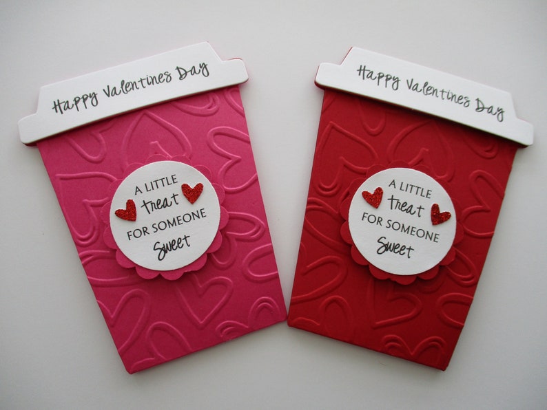 May include: Two Valentine's Day cards shaped like coffee cups. One is pink, the other red, both with embossed heart patterns. Each card has a white label with the text "A LITTLE TREAT FOR SOMEONE SWEET" and two red glitter hearts. The top reads "Happy Valentine's Day."