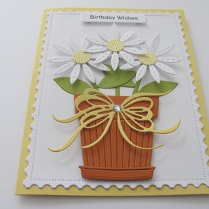 May include: A white card with a yellow border and a scalloped edge. The card has a white background with a green and brown flower pot with a yellow bow and three white flowers with yellow centers. The card says "Birthday Wishes" at the top.