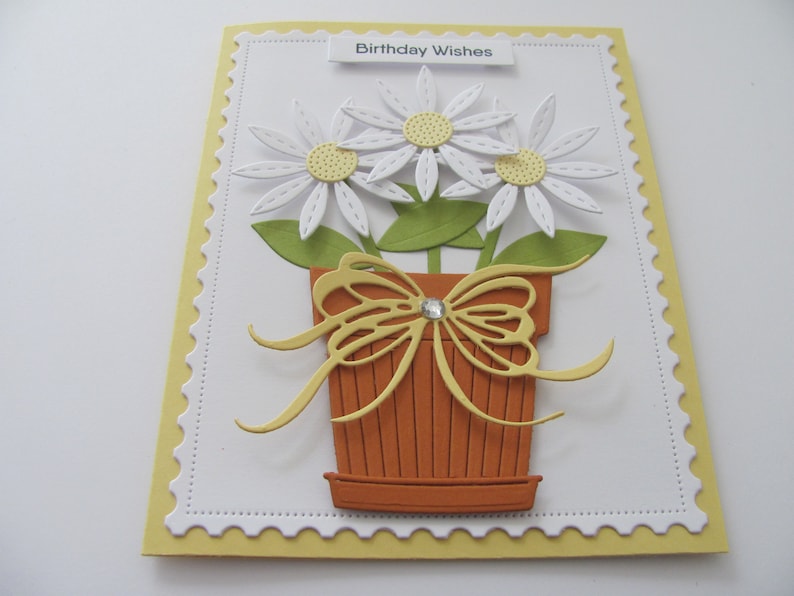 May include: A white card with a yellow border and a scalloped edge. The card has a white background with a green and brown flower pot with a yellow bow and three white flowers with yellow centers. The card says "Birthday Wishes" at the top.