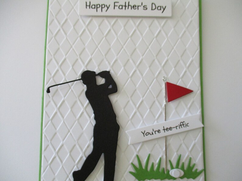 May include: A white card with a green border and a diamond pattern. The card has a black silhouette of a golfer swinging a club. The card also has a red flag, a white banner with the text "You're tee-rific", and green grass with a white golf ball.