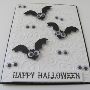 May include: A rectangular greeting card with a white embossed background featuring repeating eye patterns. Three black paper bats with googly eyes are attached to the front. Additional loose googly eyes are scattered across the card. A white label at the bottom displays the black text HAPPY HALLOWEEN.