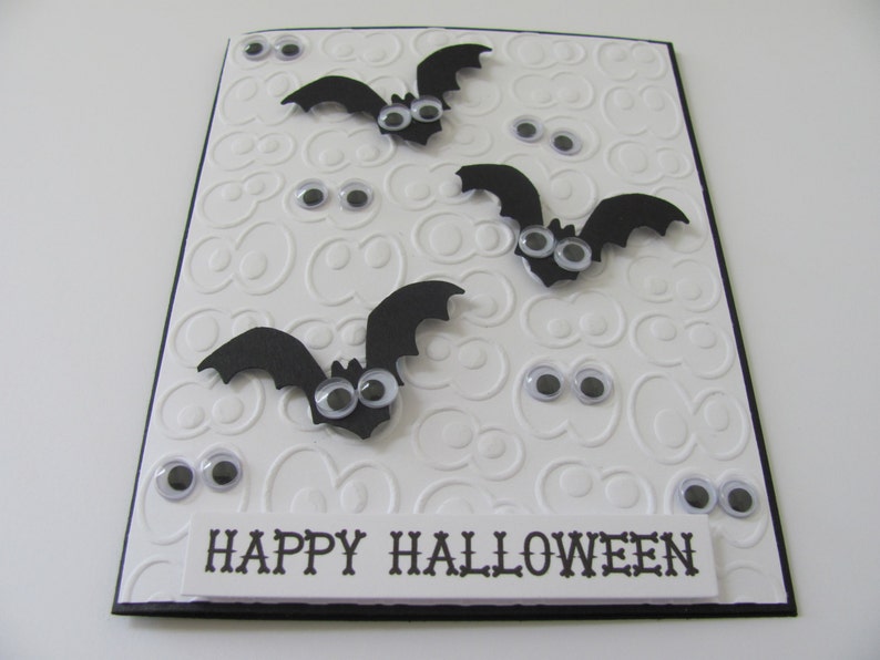 May include: A rectangular greeting card with a white embossed background featuring repeating eye patterns. Three black paper bats with googly eyes are attached to the front. Additional loose googly eyes are scattered across the card. A white label at the bottom displays the black text HAPPY HALLOWEEN.