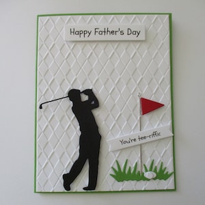 May include: A white card with a green border and a diamond pattern. The card has a black silhouette of a golfer swinging a club. The card also has a red flag and a white banner with the text "You're tee-riffic". The card says "Happy Father's Day" at the top.