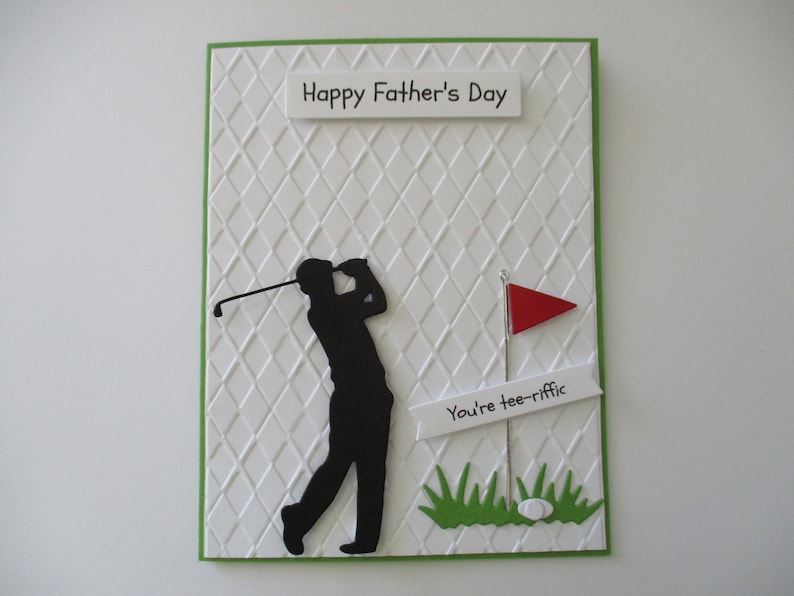 May include: A white card with a green border and a diamond pattern. The card has a black silhouette of a golfer swinging a club. The card also has a red flag and a white banner with the text "You're tee-riffic". The card says "Happy Father's Day" at the top.