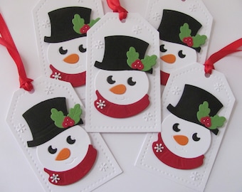 Könnte beinhalten: Fünf weiße Geschenkanhänger mit Schneemann-Gesichtern. Jeder Schneemann trägt einen schwarzen Zylinder, einen roten Schal und grüne Stechpalme mit roten Beeren. Die Anhänger haben ein weißes Schneeflockenmuster.