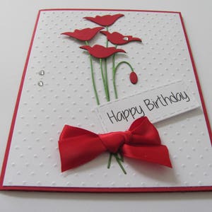 May include: A white card with a red border and red paper flowers. The card has a red bow and the text "Happy Birthday" on a white tag.