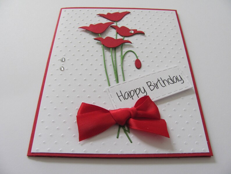 May include: A white card with a red border and red paper flowers. The card has a red bow and the text "Happy Birthday" on a white tag.