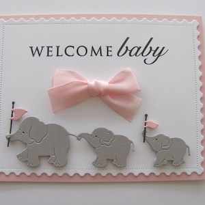 K&ouml;nnte beinhalten: Eine wei&szlig;e Karte mit rosa Rand und einer rosa Schleife. Die Karte sagt "WELCOME baby" und hat drei graue Elefanten mit rosa Flaggen.