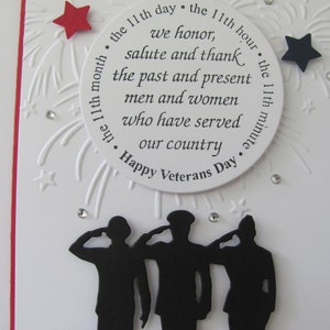 Pu&ograve; includere: Un biglietto bianco per il Veterans Day con un bordo rosso. La carta presenta un design circolare con testo in onore dei veterani, circondato da stelle e fuochi d'artificio. Sagome di soldati che salutano in basso.